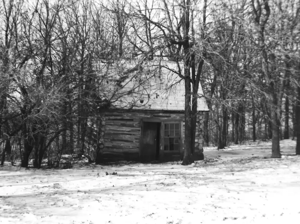 Sot-hvitt bilde av en laftet stue i skogkanten vinterstid. Nakne trær og snø på bakken. 