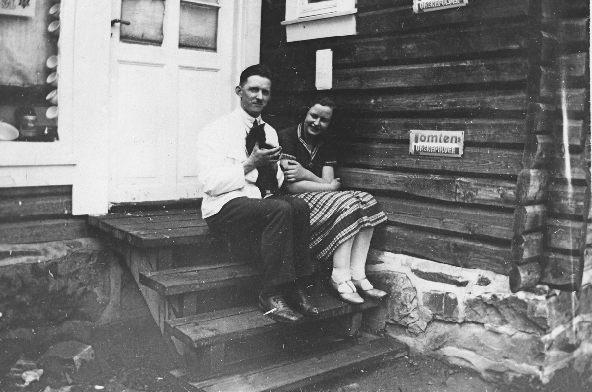 Mann med katt, og kvinne, sittende på trapp til tømmerhus.