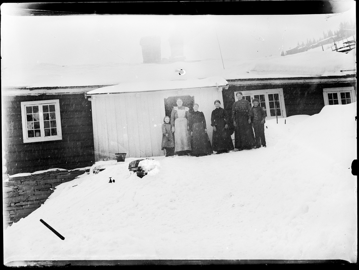 Gruppebilde av seks personer foran en husvegg, i snøvær. Fire kvinner og to barn.