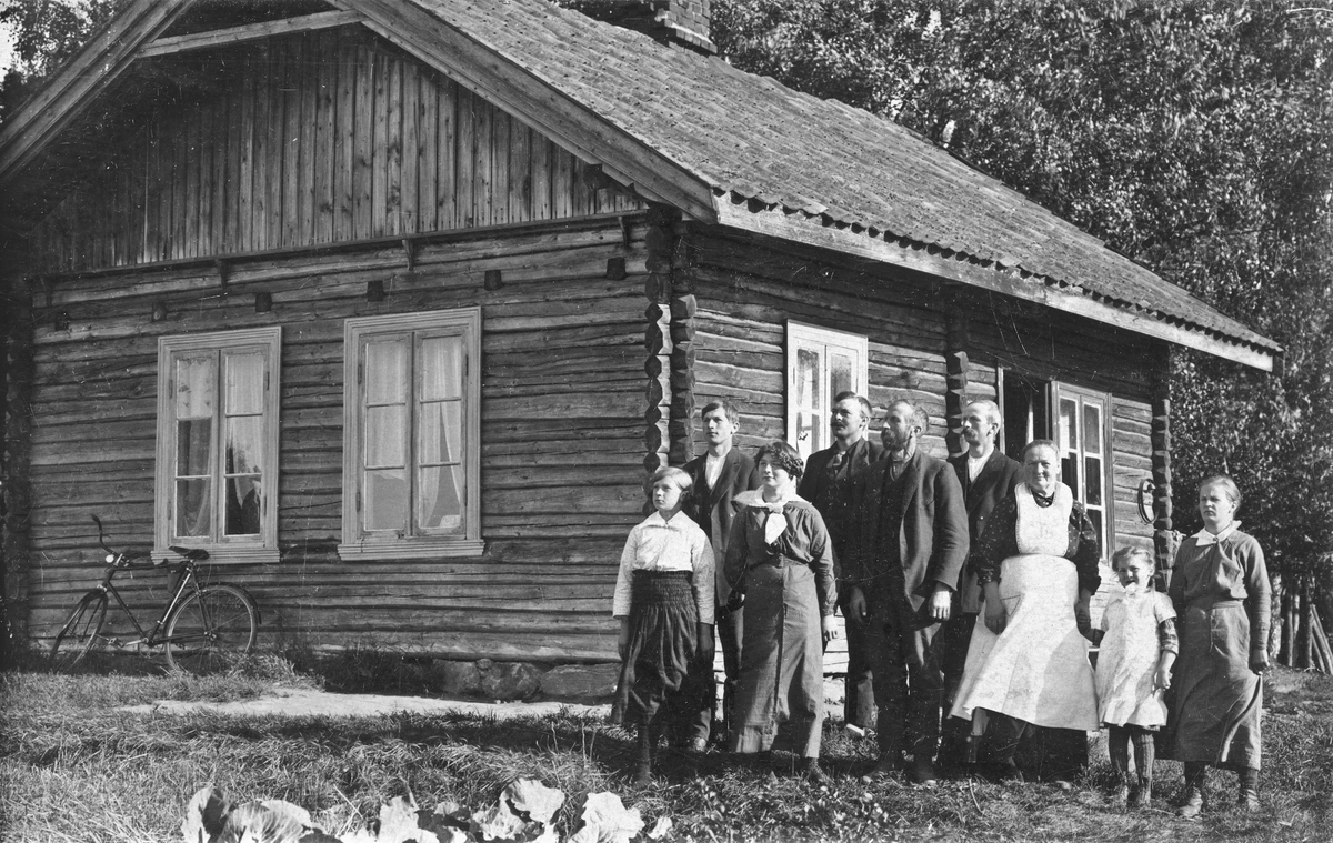 Barn og voksne utenfor et tømmerhus. Olava Vardrum.