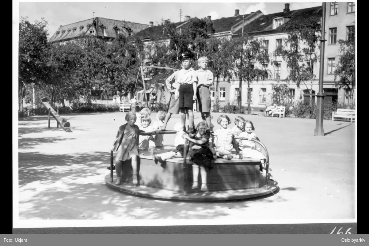 Barn i lek med karusell på plassen nederst i Thorvald Meyers gate mot Søndre gate (i bakgrunnen). Vippehuske og huskestativ. Parkbenker. (Minigolfbane idag)