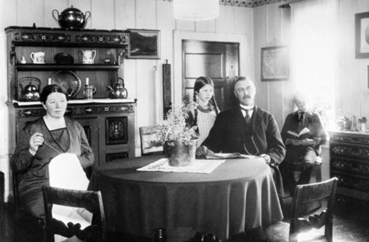 Interiør, stue, Rør store, Ringsaker. Familien Røhr rundt bordet. Mor syr, far leser avisen. Framskap, møbler.