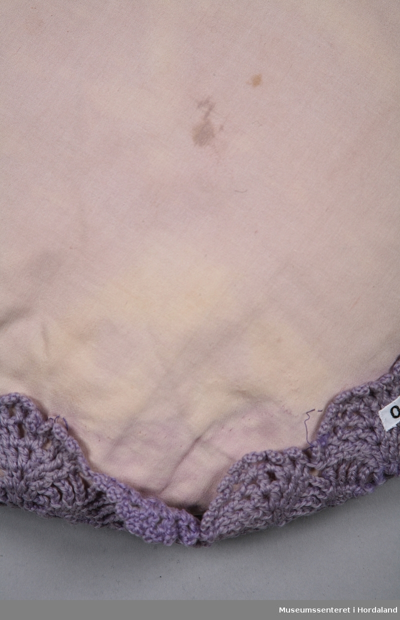 Rund hekla pute i lilla ullgarn. Det hekla framstykket er bretta rundt kanten på ei pute trekt med beige satengstoff og sydd fast.