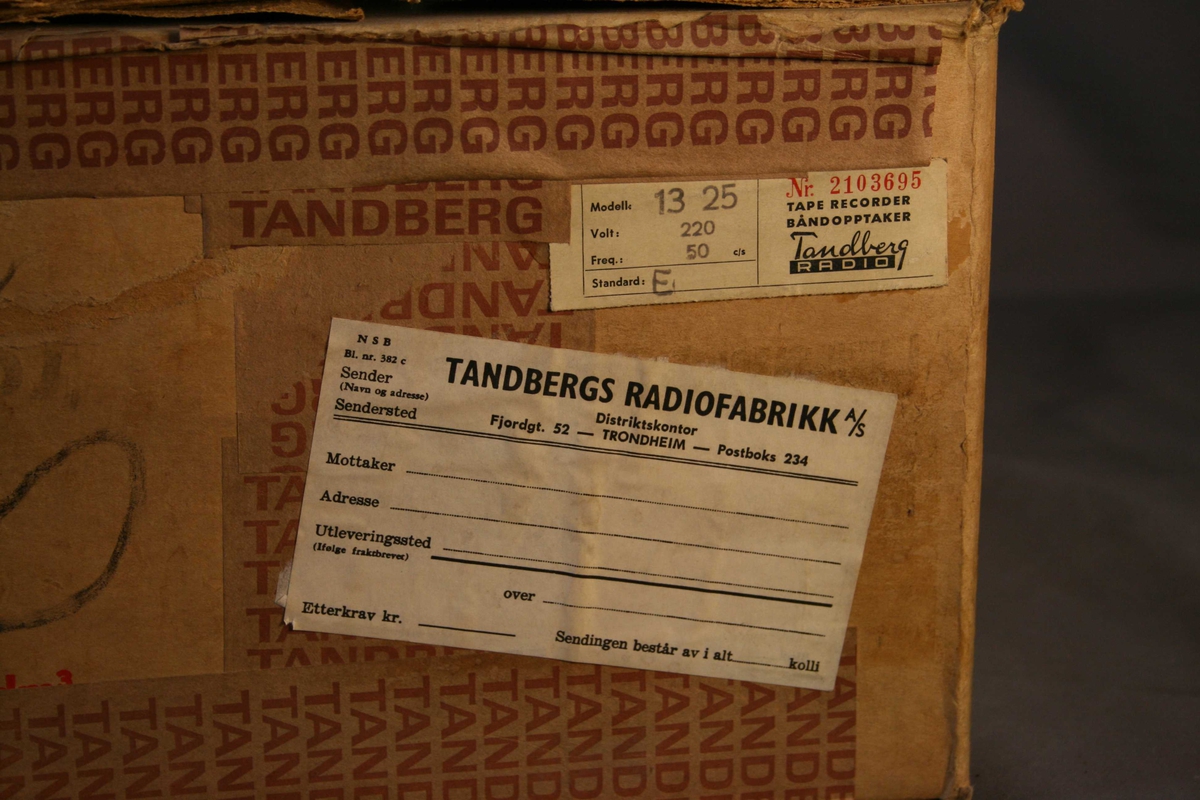 Brun rektangulær pappkasse. Tape med Tandbergs logo er festet til kassen. Isopor inne i kassen, for beskyttelse og stabilisering av båndopptakeren.
