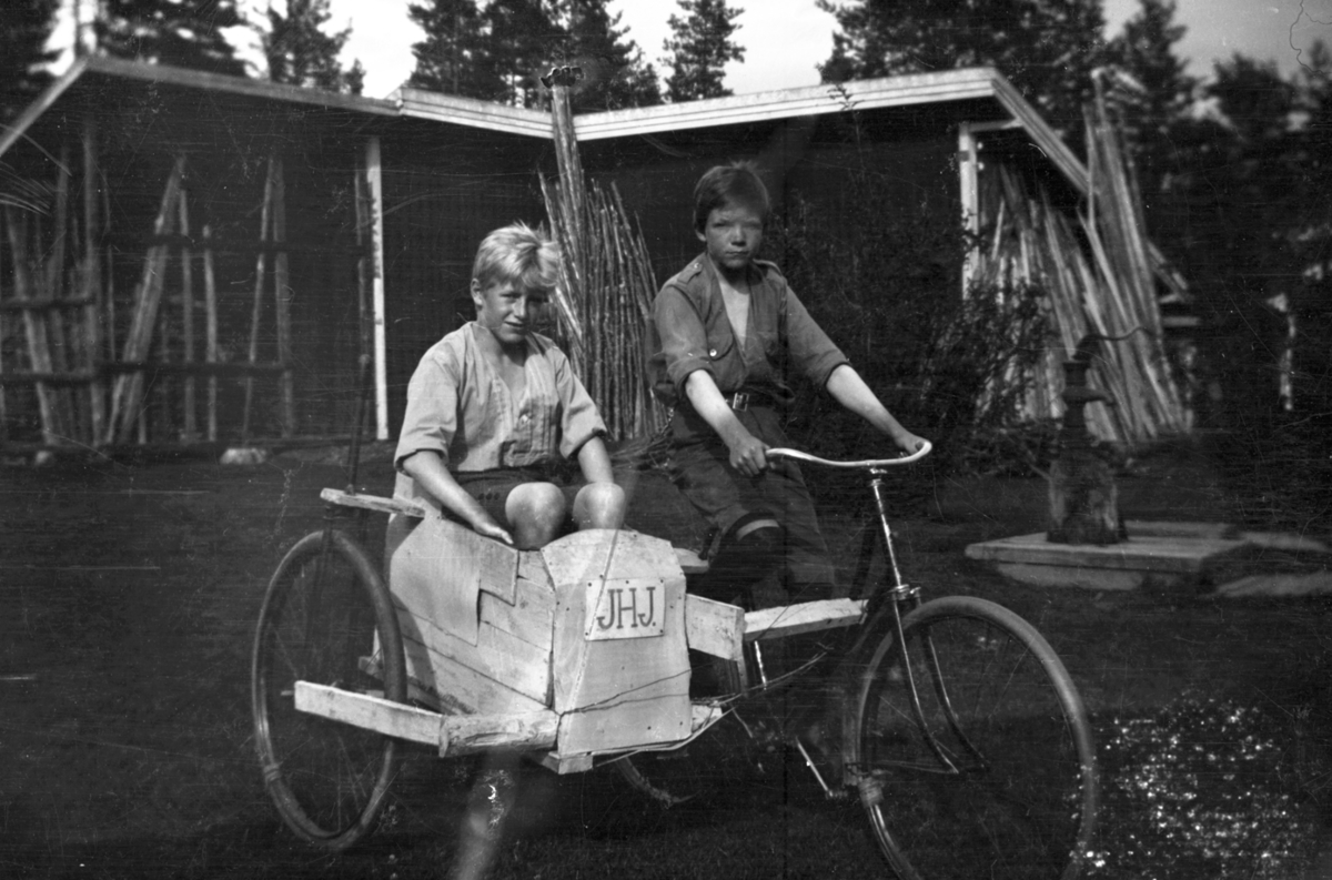 To gutter med sykkel og hjemmelaget sidevogn med skilt "JHJ". 
Vannpost bak.
Gutten til høyre er Jon Jørgensen (1908-1984), bror til Helge. Skiltet på sykkelen, "JHJ" henspeiler trolig på guttenes navn.
Gutten i "sidevogna" er Helge Jørgensen (1907-2004) fra Søbakken i Elverum. Identifiseringa er bekreftet av Helge Jørgensens sønn Anders Jørgensen.