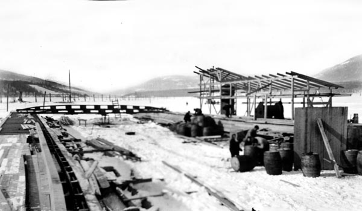 Bygging av soppeapparat ved Lillehammer lense - der Gudbrandsdalslågen renner ut i Mjøsa - vinteren 1909. Slike soppeapparater (enkelte steder også kalt «mosemaskiner») besto av to pongtonger med flåteganger, som var forbundet med tverrstilte bruer. Den nedre brua var faststående, den øvre var bevegelig. Begge hadde nedfellbare stenger («bein»), som ble brukt til å presse sammen det løstømmeret som befant seg inne i soppeapparatet.  Da dette fotografiet ble tatt, var innretningen uferdig. Den lå på ei snødekt isflate på Vingneslandet.  Isen ble tydeligvis brukt som arbeidsplattform. Pongtongene med gangbaner og skinnegang var langt på veg ferdige, men skuret der den motoriserte vinsjen som skulle drive anlegget skulle stå, var fortsatt bare et bindingsverksskjellett. Når soppeapparatet var ferdig skulle det plasseres i den nedre delen av lenseanlegget, der ferdigsortert tømmer ble ledet med den svake strømmen inn mot soppeapparatet, der den øvre brua sto med oppslåtte «bein», slik at stokkene la seg parallelt mot den nedre brua, hvor beina var felt ned i vannet. Når soppeapparatet var passe fullt, felte lensekarene ned «beina» på den øvre brua, og kjørte den, med vinsjen som trekkraft, mot den nedre brua.  Dermed ble tømmeret presset ihop, slik at det kunne slås vaiere rundt «soppa» - tømmerbunten. Deretter hevet de «beina» på den nedre, faststående brua, slik at tømmeret kunne flyte ut av anlegget. Soppene ble samlet opp og bundet sammen i «slep», som ble buksert sørover Mjøsa. Før dette kunne skje måtte tømmeret være sortert, slik at det tømmeret som skulle til sagbruk og treforedlingsindustri i Mjøs-regionen kunne skilles fra det som skulle fløtes videre nedover Vorma og Glomma.

Da Kristiania Tømmerdirektion i 1903 vedtok å overta administrasjonen av fløtinga på Mjøsa hadde Lillehammer lense lenge hatt lokalt eierskap. Den nevnte beslutningen fikk i første omgang ingen annen konsekvens for lensedriften enn at det fra da av var Tømmerdirektionen som inviterte til anbud på driften av de 8 lenseanleggene som fantes der fløtingsvassdragene rant inn i innsjøen. I 1908 inngikk Kristiania Tømmerdireksjon forpaktningsavtale for Lillehammer lense, og det var denne organisasjonen som moderniserte anlegget. De to første sesongene, i 1909 og 1910, ble soppeapparatene drevet av en dampmaskin. Etter dette inngikk Tømmerdireksjonen avtale med Lillehammer kommunale elektricitetsverk om leveranser av elektrisk energi. Fra da av ble soppinga av tømmer utført med elektromotorer som drivkraft. Trepongtongene vi ser på dette fotografiet ble erstattet av stålpongtonger fra Hamar Jernstøperi og mek. Verksted våren 1916.

Onsdag 12. mai 1909 beskrev avisa «Lillehammer Tilskuer» det nye soppeapparatet ved byens lenseanlegg slik:

«Soppemaskinen ved Lændsen er nu paa det nærmeste færdig. Det bestaar av to 64 Meter lange flydende Broer, der holdes sammen av to 12 M. lange Tverbroer. Paa den ene Langside ligger Maskinen. Hele Apparatet ligger paa langs i Strømretningen. Tømmeret kommer drivende ind i den øvre Ende, og føres af Strømmen ned mod to flere Meter dybe Arme, der stikker ned fra den nederste Tverbro. Her hobes det da op, og det slig, at Stokkene blir liggende paa tvers imellem begge Langbroer. Naar hele Rummet er fyldt trækkes en forskyvelig Bro, der ogsaa er forsynet med nedstaaende Arme, nedover af Maskinen, og derved presses Tømmeret sammen i en flere Meter dyb Sop. Der slaaes Staaltraadbaand om, og Soppen er færdig. Armene paa den nederste Tverbro løftes op og Soppen flyder ud. Saa begyndes der paa igjen paa samme Maade.»

Dampmaskinen som drev anlegget fra starten av kunne fyres med ved.