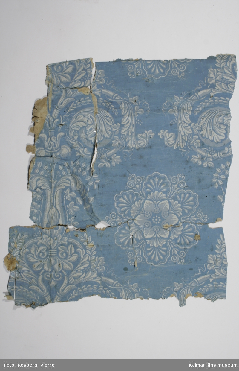 KLM 22998:1. Tapet, av papper. Tapet med blomsteruppställningar i ljust blått och grått tryckt på blå botten. Mellan blomsteruppställningarna stora blommor. På baksidan fragment av träflisor. Datering: 1840-tal.