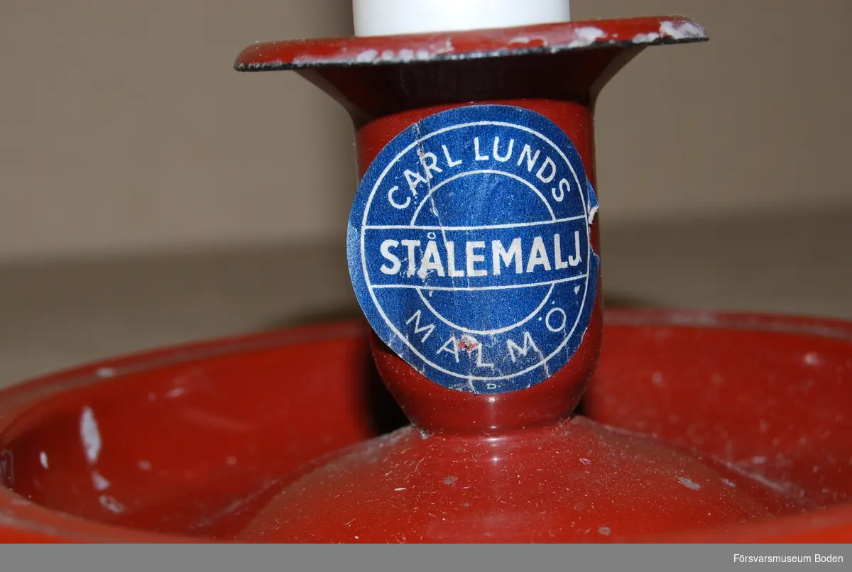 Ljusstakar av järnplåt med röd emalj från Carl Lunds Stålemalj, Malmö. Etikett med texten Cm.14 på undersidan, vilket troligen är modellbeteckningen. Oanvända stearinljus medföljer.