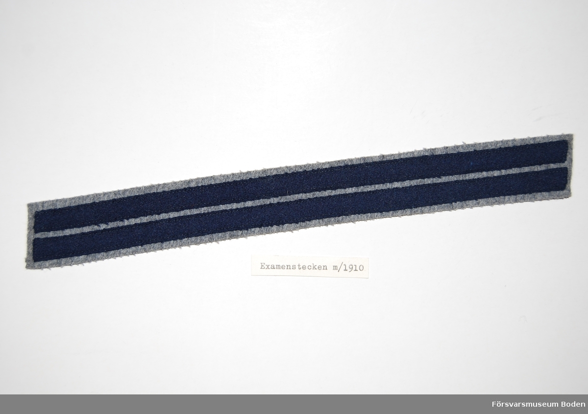 Två smala band av blått kläde på bakgrund av grått kommisskläde. Sys fast på vapenrockens högra överärm. För modellexemplar, se AM.015023.