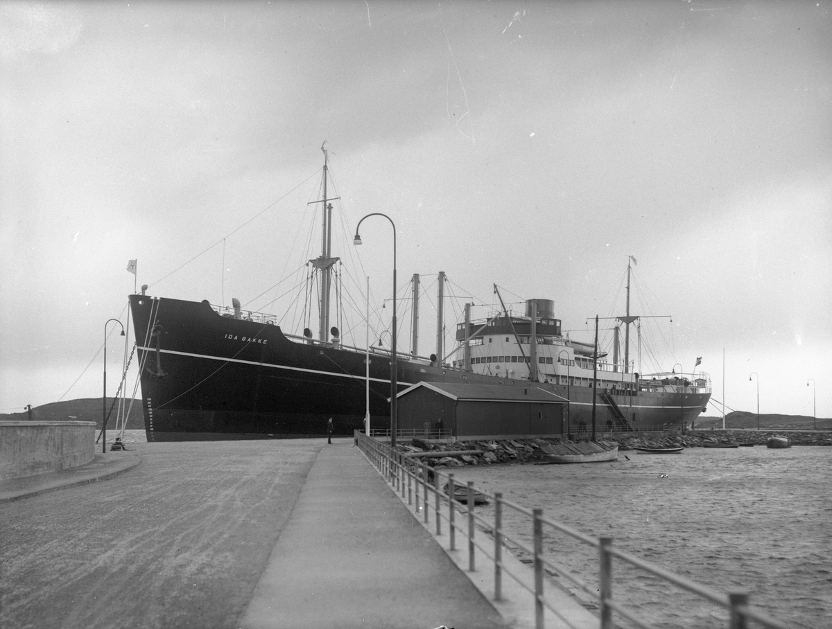 Linjeskipet "Ida Bakke" ankret opp. To mindre båter i forgrunnen foran skipet. Fjell i bakgrunnen.