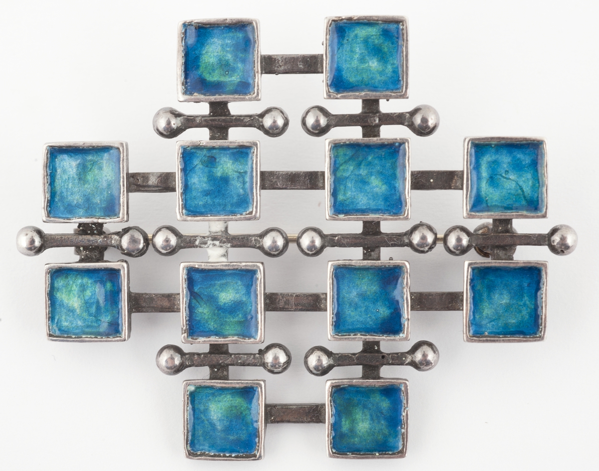 Brosje i støpt sølv med tolv kvadratiske elementer fylt med emalje i blågrønne sjatteringer. 