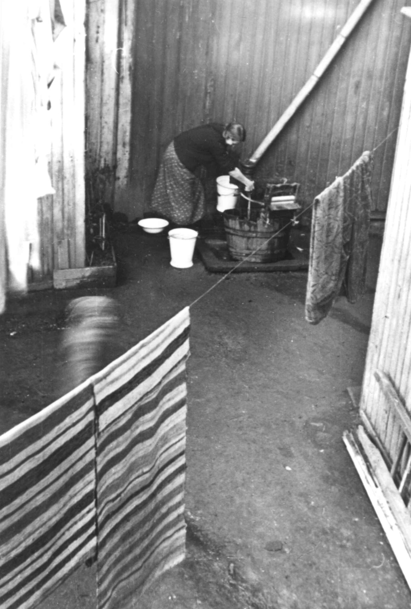Klesvask, kvinne vasker tøy utendørs, utstyrt med vannpost, rulle/maskin til å presse ut vann, emaljerte bøtter og fat. Filleryer til tørk på snor. Jfr. NF.13400-072.
Fra boliginspektør Nanna Brochs boligundersøkelser i Oslo 1920-årene.