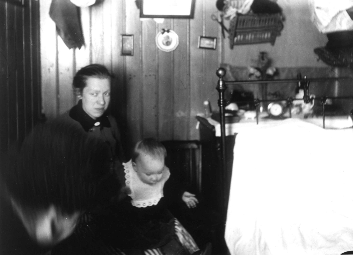 Interiør, Oslo. Kvinne og barn sitter ved seng i tett møblert rom.
Fra boliginspektør Nanna Brochs boligundersøkelser i Oslo 1920-årene.