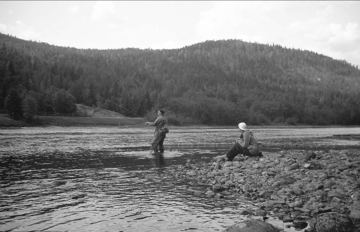 Daghild Skapell Jakobsen sitter ved elvebredden av Begna og ser på en mann som fisker.