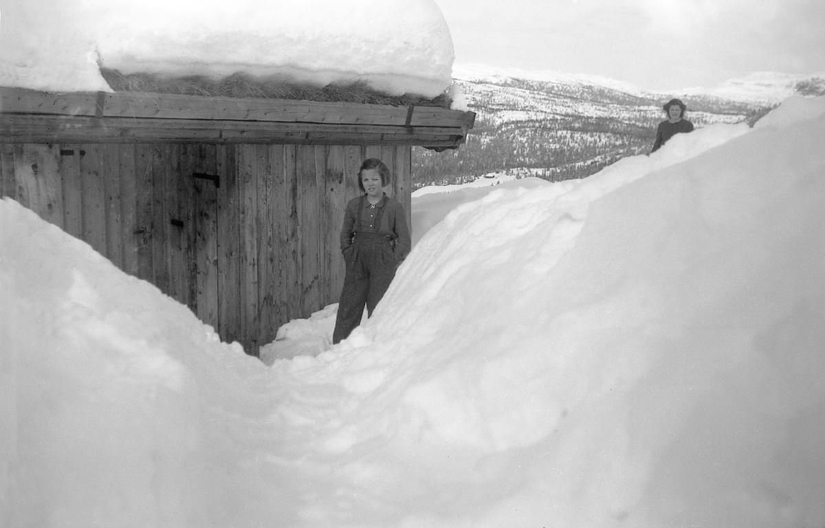 En jente står mot en vegg, masse snø rundt, Ligardshaugen. Fotografert 1947.