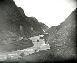 Smal elvedal med gårdsbruk, bro over elven og foss innerst i