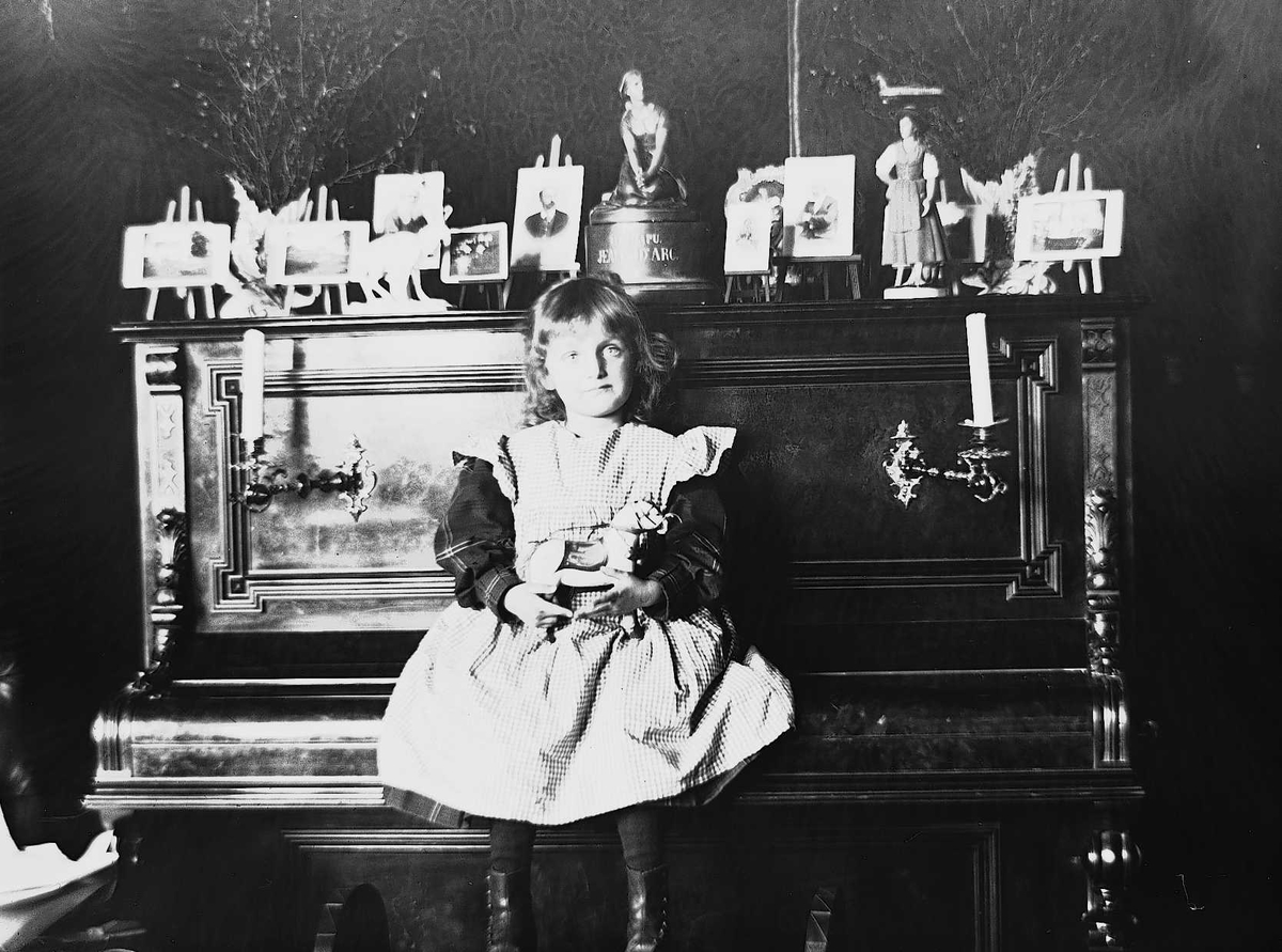 Karen Q. Wiborg sitter på piano i stue, Meltzers gate 9, Oslo, 1899. Fotografier i ramme plassert på pianoet.