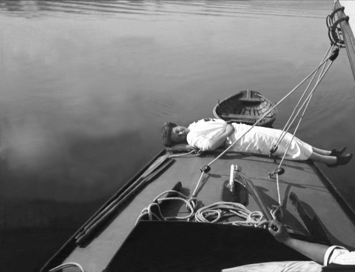 En kvinne ligger på dekk og soler seg, en person styrer seilbåten. Robsahm og Lund.