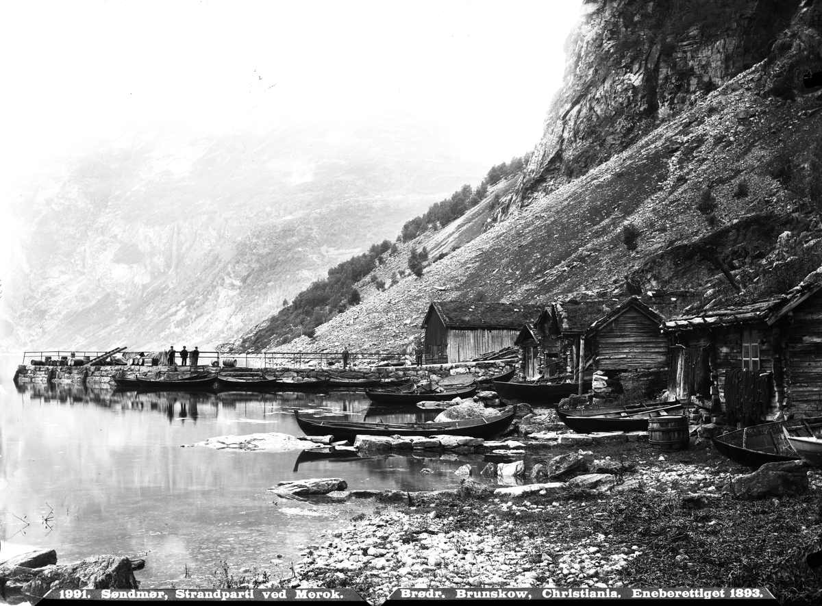 Strandparti med båter, naust, tønner og brygge i Merok ved Geiranger. Fotografert av Brødrene Brunskow i 1893 med eneberettigelse.