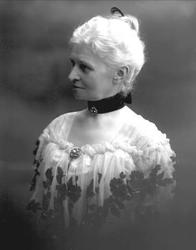 Portrett, eldre kvinne hvit, blomstrete kjole. Halssmykke på