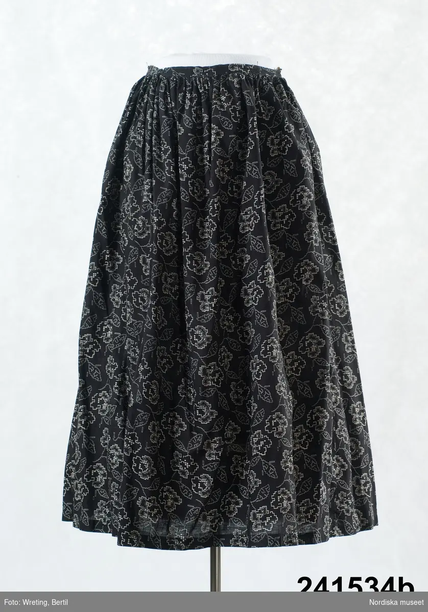 2-delad klänning bestående av liv och kjol a-b. Av bomullslärft svart med tryckt vitt stiliserat blommönster.
a. Klänningsliv sytt som en blus med 2 framstycken/sidstycken, rynkade mot besparing. Ett ryggstycke rynkat i midjan mot ett fatsytt smalt band av tyget som knyts fram.  Rak krage, knäppning  med 5 tryckknappar av gulmetall Isydd ärm  med en söm, tyget skarvat på ärmens utsida med ett tryckt rutigt tyg i svart och vitt. ärmen rynkad mot 1,5 cm bred ärmlinning knäppt med tryckknapp. Fodrad i övre delen och i ärmarna med ljusgrå bomullslärft, fodret knäpps med lösa framkanter med 3 par hakar och hyskor. Maskinsydd
Anm. Sliten på ärmlinningarna, ett par små hål mitt fram, något blekt.
b. Kjol i 3 våder, rynkad mot smal midjelinning av tyget, Sprund knäppt med 2 hakar och hyskor och en tryckknapp. 
Har tillhört Augusta Davidsson, "Snögges Augusta" f. 1868 i Habo död 1950 på ålderdomshemmet. Hon hade bott hela sitt liv i föräldrahemmet ett soldattorp under Vagnhult i Habo. hennes far var soldat liksom brodern Efraim. Först vårdade hon modern i hemmet, senare hushållade hon åt den ogifte brodern Efraim. Sen gick hon på "hjälpe" hos bönderna i bygden. Foto av stugan skall finnas i museet. Kläderna som museet mottagit är de som hon hade med sig till ålderdomshemmet.
/Berit Eldvik 2011-01-17