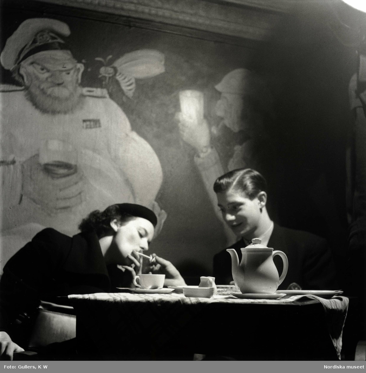 Ett ungt par vid ett cafébord. Mannen tänder kvinnans cigarett.