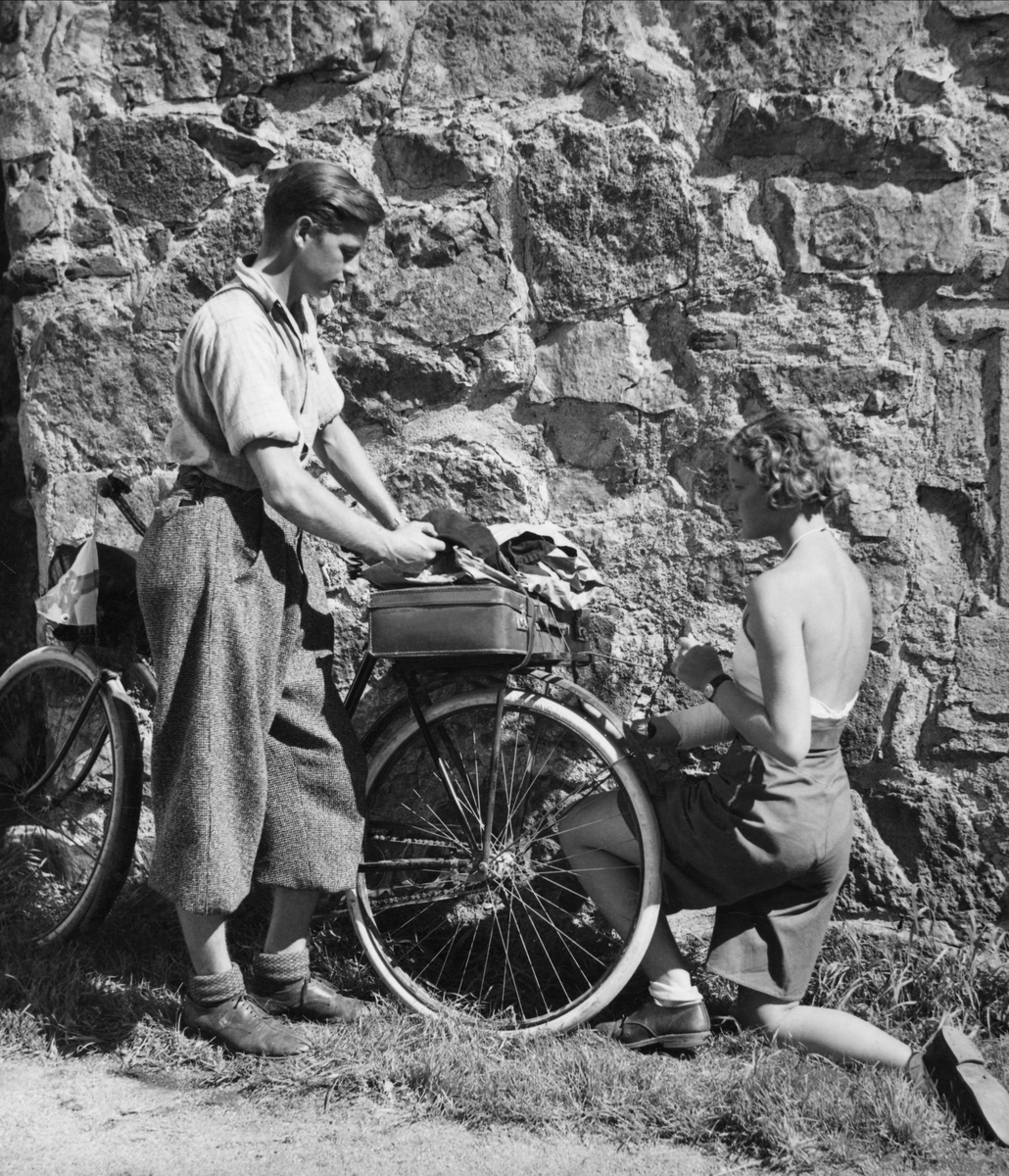 Två cyklister, en ung man och en ung kvinna, har stannat vid en stenmur. han är klädd i vida knäbyxor och skjorta, hon har kortbyxor och ett liv med halterneck.