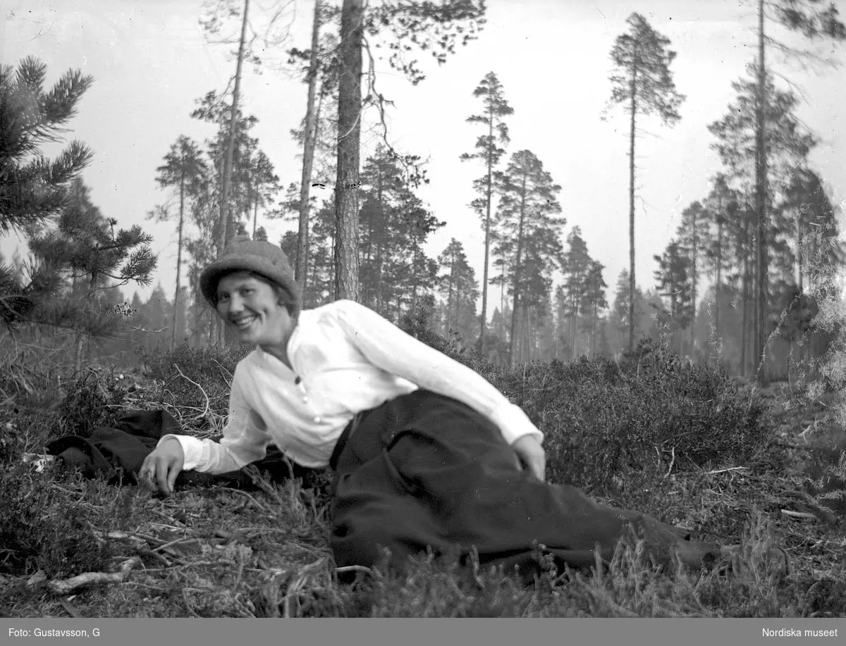 En kvinna iklädd ljus skjorta, mörk kjol  och hatt ligger i skogsbacke.