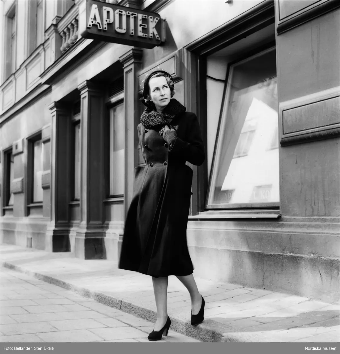 Modebild för Sahlins konfektionsfabrik. Kvinna iklädd kappa och högklackat och hatt  kliver ned från trottoaren utanför apoteket.