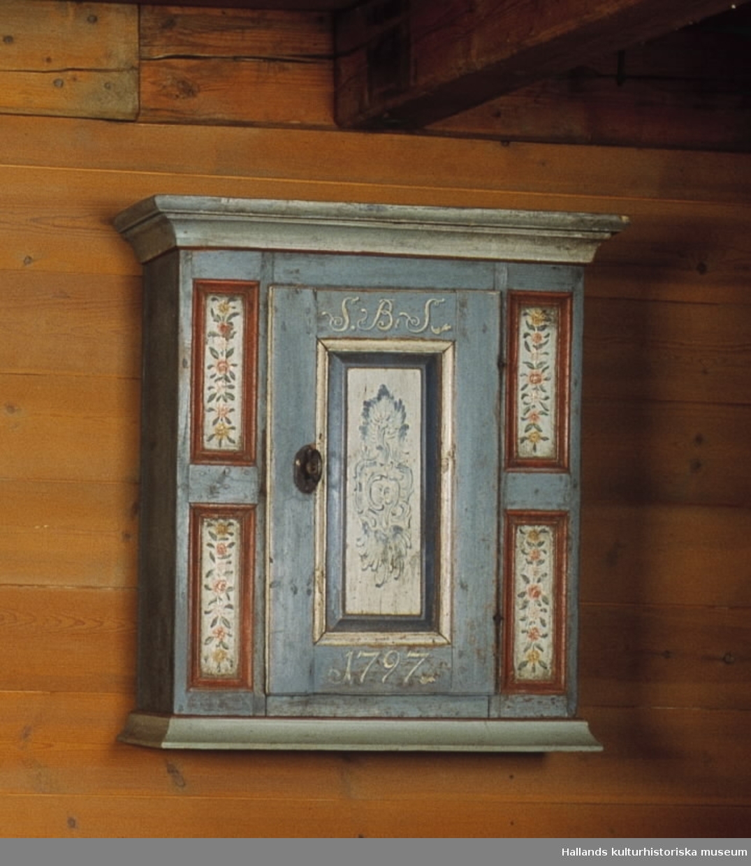 Väggskåp, blåmålat med rocaille på dörren. Märkt SBS 1797. En hylla inuti.