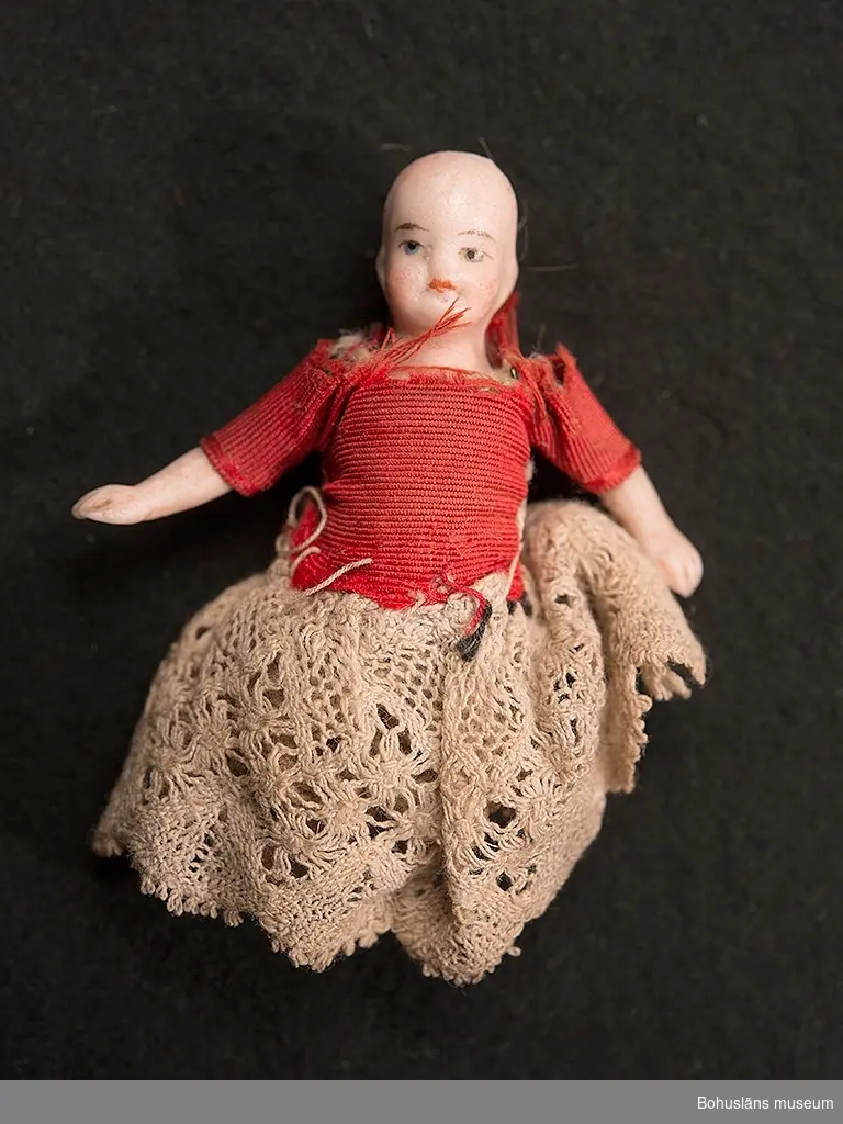 Liten docka i porslin med ledade ben och armar. Spetskjol och blus i röd ripssiden, virkad spetsunderkjol. Dockans ben är avbrutna.
Dockskåpsdocka.