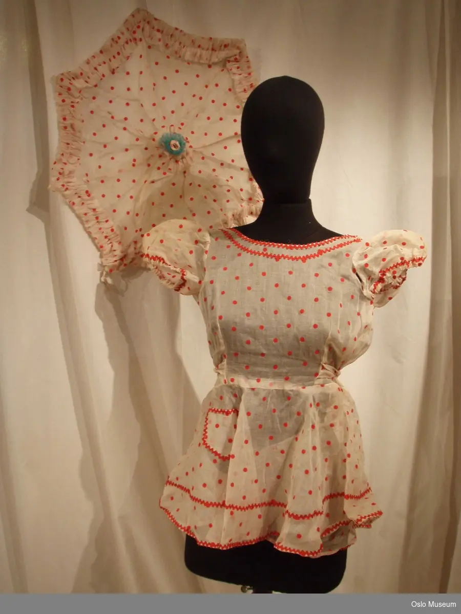 Kort ballettkjole, truse og parasoll (kun trekket) i hvitt transparent stoff med røde prikker. Røde sikksakkbånd på kjole.