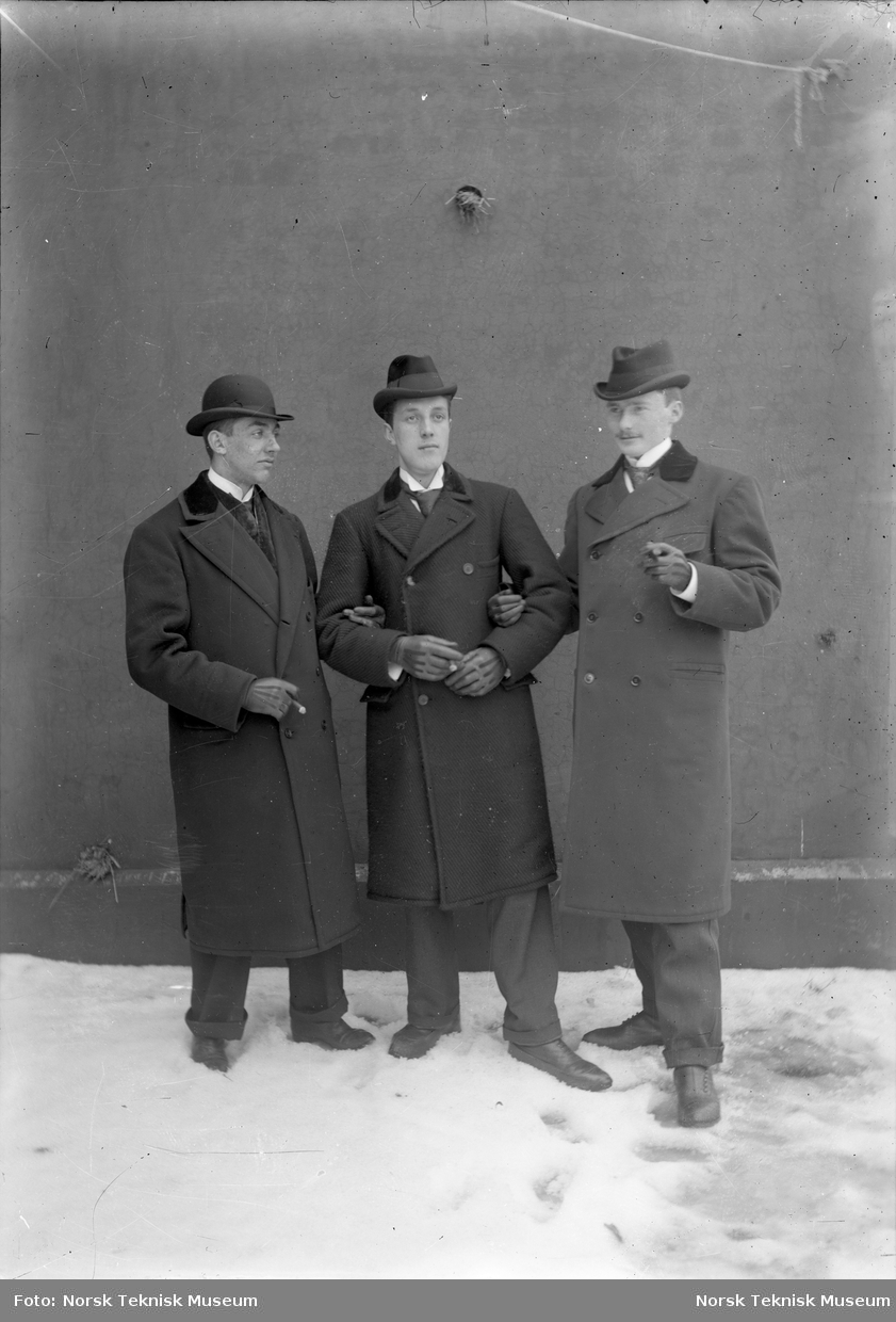Tre menn med sigarer, hatter og frakker poserer arm i arm foran en vegg .