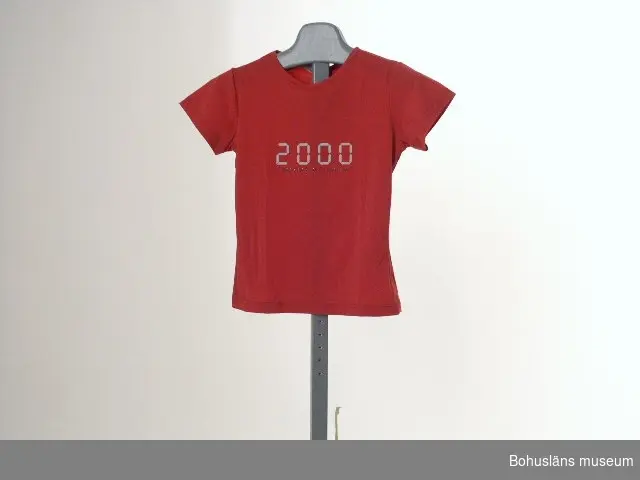 Rosa t-shirt i töjbart konstmaterial med påtryck "2000" i grått samt "MESSAGE MILLENNIUM" i silverglitter,  inköpt i Uddevalla för 299 kronor.
Storlek small. Plagget märkt med två vidhängande pappersetiketter med skötselanvisning.

För information om Millennieinsamlingen, se UM2736