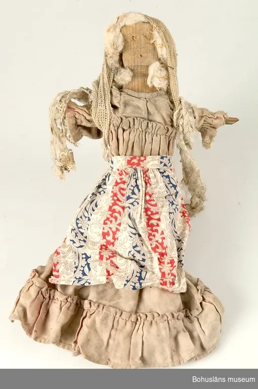 Hemmagjord trädocka från tidigt 1900-tal. Dockkroppen är grovt utskuren ur ett stycke furu och har ansikte med antydan till mun, näsa och ögon, påspikade armar som står rakt ut utan händer och raka ben utan fötter. Hår i form av bomull. Dockan är klädd i  en mycket blekt rosa klänning av sekelskiftessnitt med volang och garnering och med huvud- och axelschal av vit spets.
Klänningen verkar ha hört till en annan, finare dockkropp.

Enl. givaren kan dockan ha tillhört Ingeborg Hammar i Dragsmark,  f. 1916. Huset där dockan är funnen är byggt 1916, Klostergård 3, Dragsmark. 
Dockan är funnen i huset sommaren 2000 under en golvbräda på  vinden.