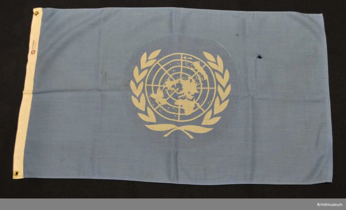 Blå flagga med vitt FN-emblem. Med kulhål i ena hörnet.