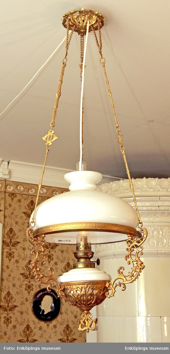 Fotogenlampa med kupa i vitt överfångsglas, hållare och tre stycken kedjor i genombrutet mönster av förgylld formgjuten metall. Fotogenhus av vitt porslin, ofärgat lamoglas. Tillverkad i slutet av 1800- talet.