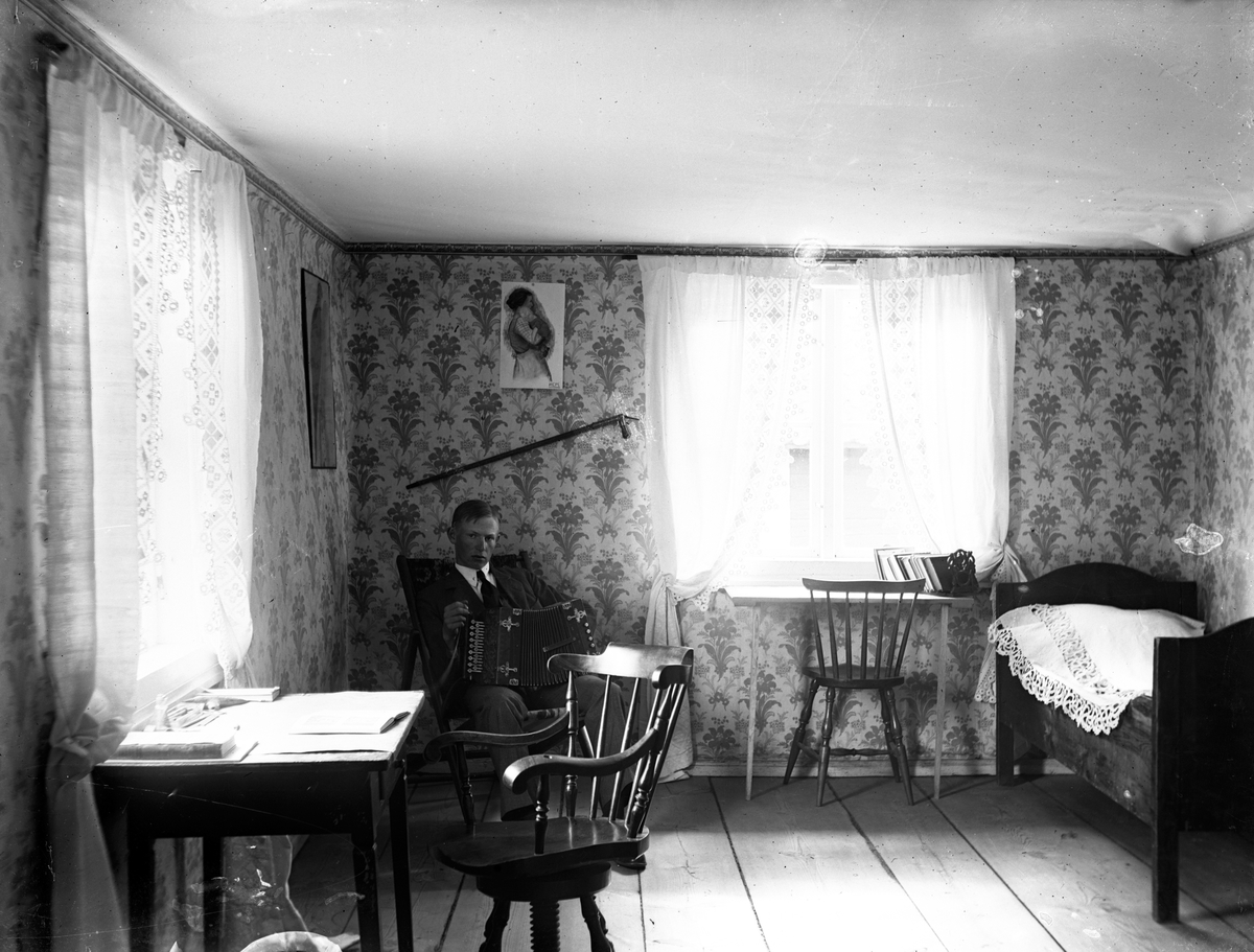 I ett rum med breda golvplankor och blommig tapet sitter en ung man med ett dragspel i knät. I andra hörnet står en säng täckt med ett vitt överkast. Vid ena fönstret står ett bord och en snurrstol i trä. Båda fönstren har vita spetsgardiner.
