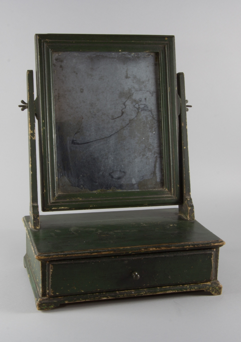 Toalettspegel av trä, grönmålad, med kvadratisk spegel löst monterad med muttrar vid två raka ståndare och med låda med knopp.