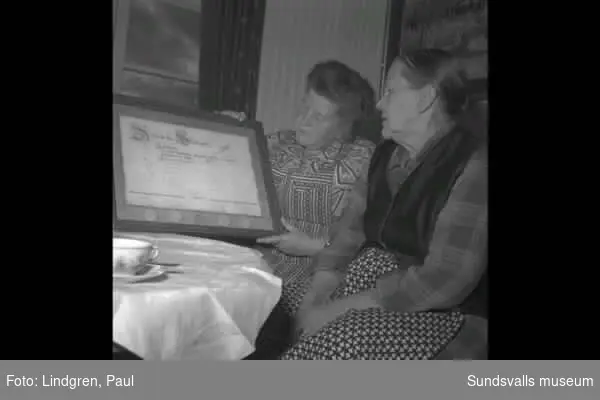 Två kvinnor från Storhullsjön, sitter vid ett bord och håller en tavla, ev diplom. Enl uppgift heter kvinnan till vänster Märta Larsson.