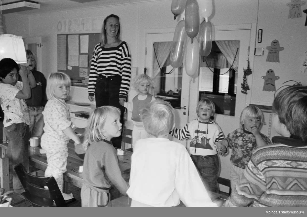 Flertalet barn och förskollärare Britt Johansson står på stolarna runt ett bord i köket. Några håller varandra i händerna. På bordet finns kaffekoppar och fat med bakverk/tårta på. I taket hänger uppblåsta ballonger. På väggen bakom hänger uppklistrade Spöket Laban-figurer som barnen har gjort. Hoppetossan, Katrinebergs daghem 1992.