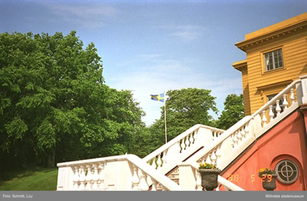 En rödokra-målad trappa med vitt räcke av sten som leder upp till Gunnebo slott. Man skymtar en liten del av slottet strax ovanför trappan. I bakgrunden ser man en flaggstång med Svenska flaggan hissad.