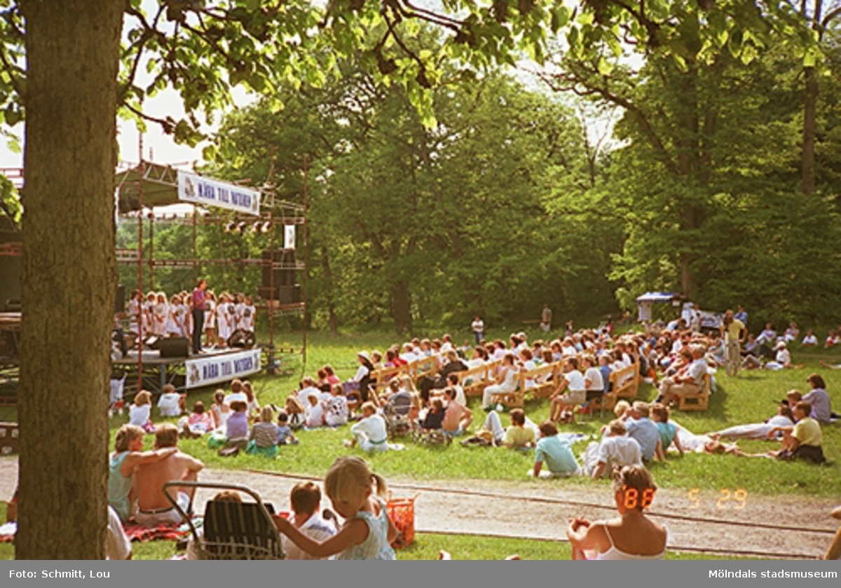 Människor som sitter i gräset och lyssnar på Lasse Berghagen som sjunger på en tillfälligt uppbygd scen. Gunnebo slotts park, 29 maj 1988.