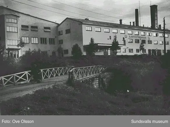 Text: "Ute vid Nacksta återfinns Nedre Norrlands Slakteriförenings kontors och fabriksbyggnad". I förgrunden bron över Sidsjöbäcken vid dess utlopp i Selångersån.