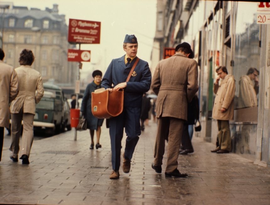 Brevbäraren är iklädd uniform av modell 1974. Brevbärarväskan var en populär modell som användes in på 1980-talet. På postväskan syns 1965 års postsymbol.
