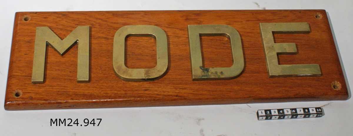 Fernissad träplatta. På framsidan bokstäverna MODE i guld. På långsidorna sitter fästbyglar i metall, två på varje.