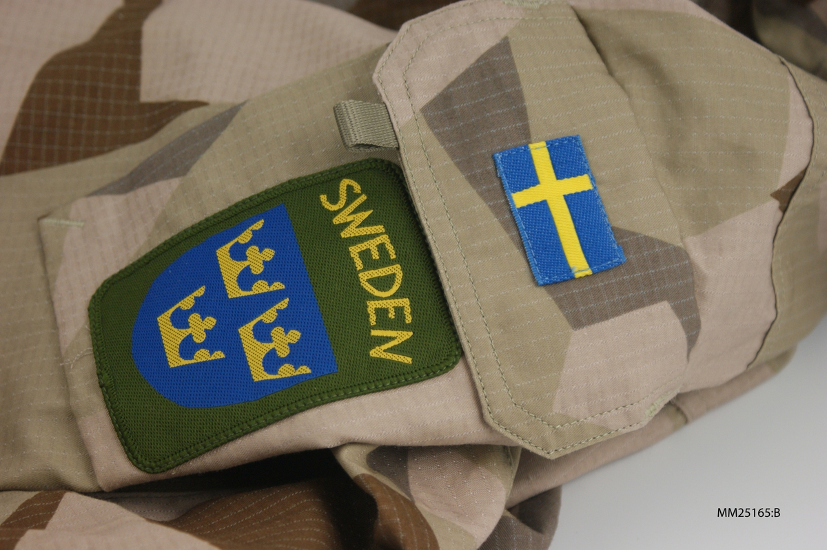 Fältjacka i beige, brun och ljust grön kamouflage. Knäpps med dragkedja som kan täckas över med en flärp med tryckknappar. Jackan har kapuschong med förstärkta kanter. På vänster ärm sitter en svensk flagga och ett märka med tre kronor och text "SWEDEN".