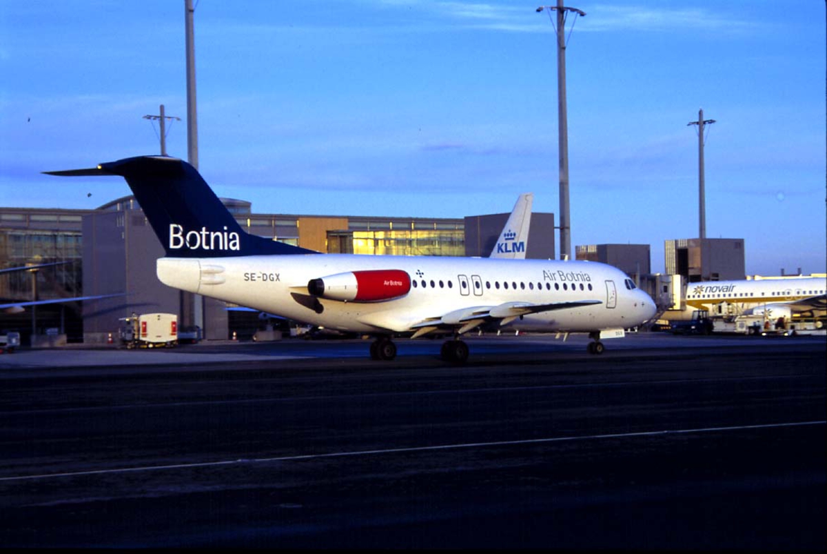 Lufthavn, 1 fly på bakken, 1 Fokker F28-4000 Fellowship SE-DGX "Vemund Viking" fra Air Botnia, sett fra siden. Terminalen med flere i fly i bakgrunnen.