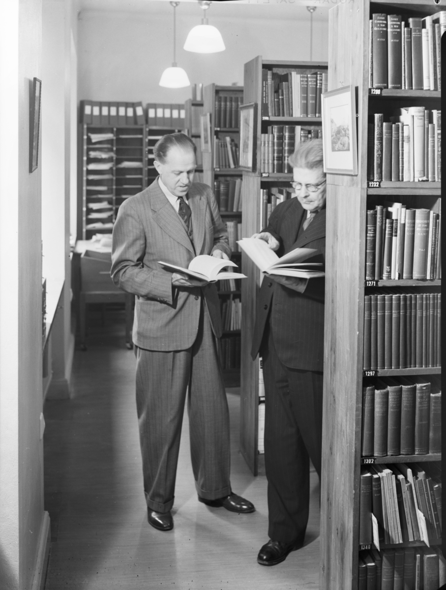 Två män stående med vasin bok i ett bibliotek
Interiör