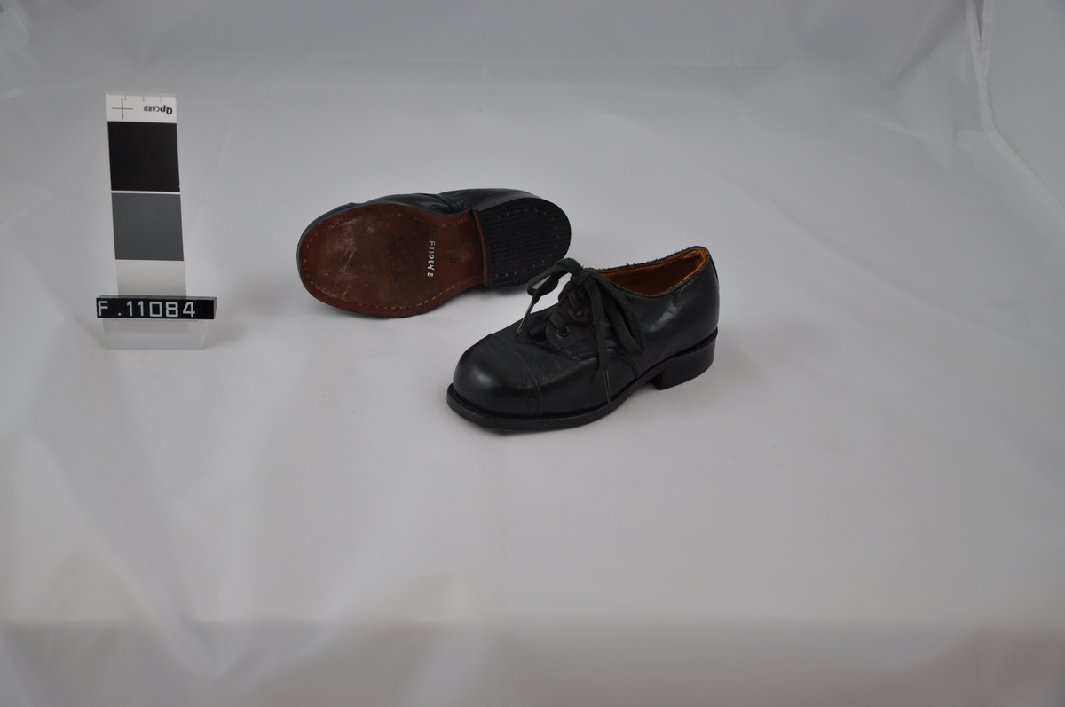 Barnesko med rett hæl og snøring. Skoen er sydd, såle limt og hælen stiftet. Bånd med hullmønster tvers over skoen mot tupp.